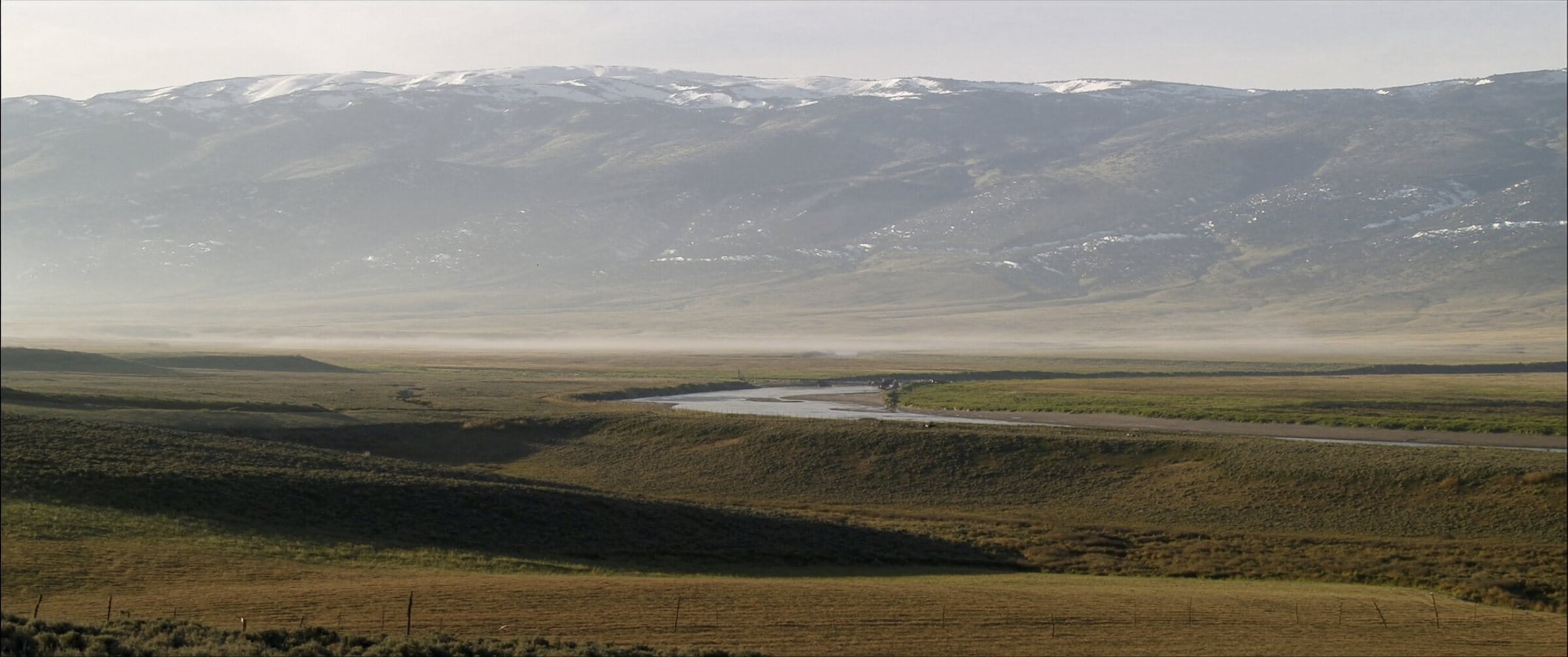 A wide Western ranch valley at first light, sage benches running up to a snow-dusted range.